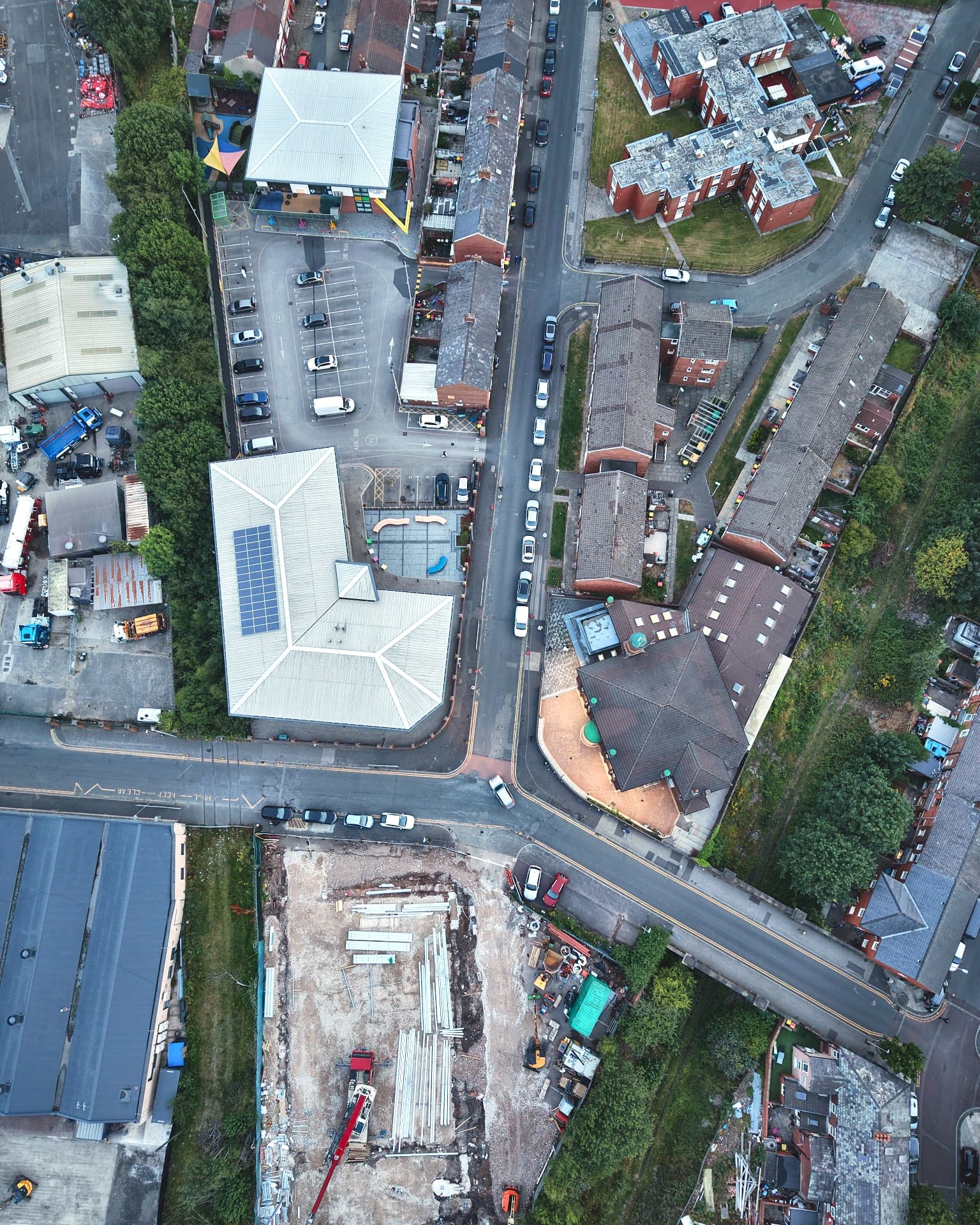 Near-vertical aerial photograph of the Quwwat campus in Deepdale, Preston, in portrait. Reading down the frame: the nursery and its play area, the car park, the Education Centre with its solar-panelled roof, Quwwatul Islam Masjid across the road to the right, and the Quwwat Academy site under construction at the foot of the frame.