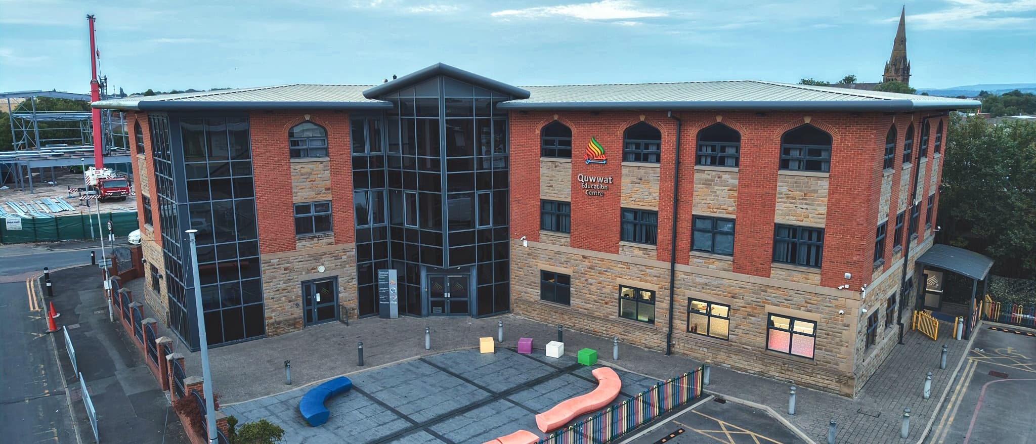 The Quwwat Education Centre on a bright evening — a three-storey red brick and stone building with a full-height glazed entrance tower, the Quwwat Education Centre sign on the front elevation, and a play area in the forecourt.