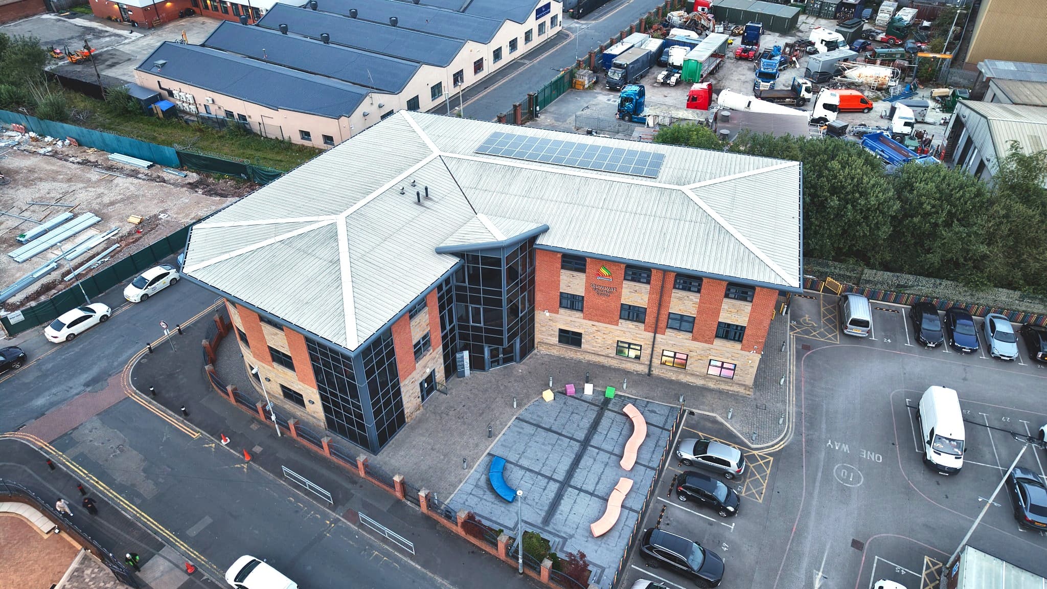 The Quwwat Education Centre from above at an angle — solar panels across its roof, the forecourt play area, the car park, and the Academy site under construction beyond.